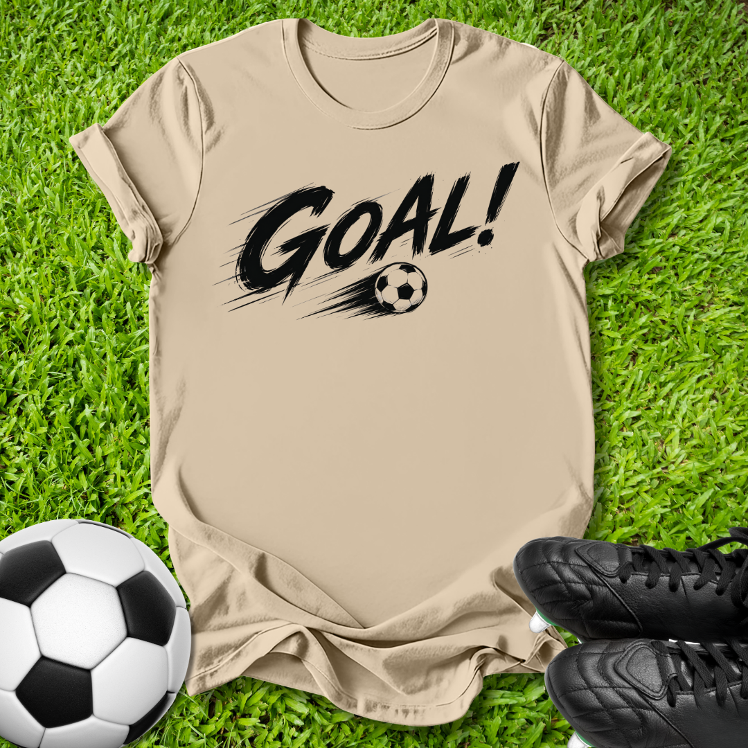 A beige short-sleeve t-shirt with a black 'GOAL!' graphic and soccer ball print, laid flat on green grass with a soccer ball and black cleats as props.