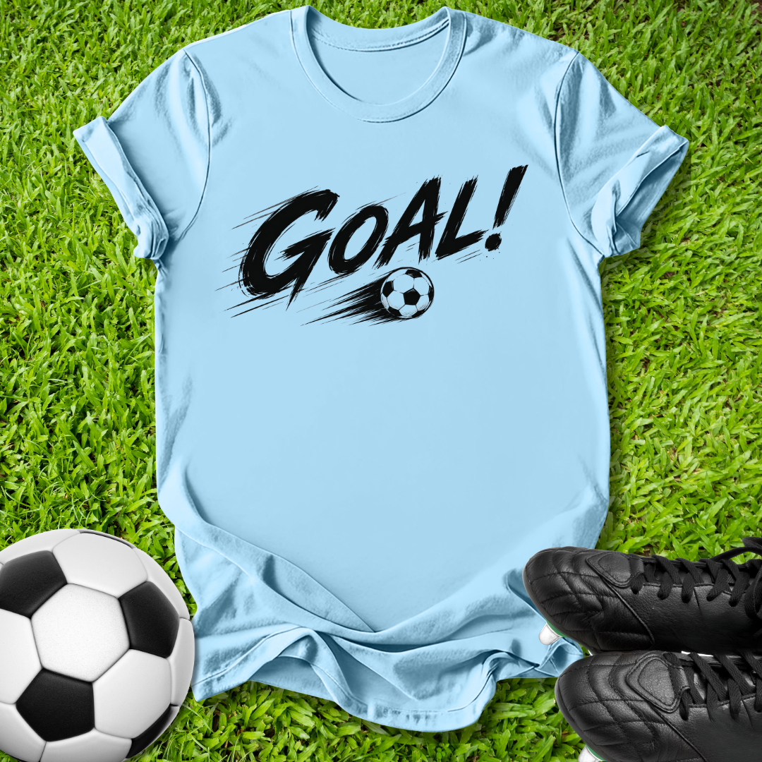 A light blue short-sleeve t-shirt with a black 'GOAL!' graphic and soccer ball design, laid flat on green grass with a soccer ball and black cleats nearby.