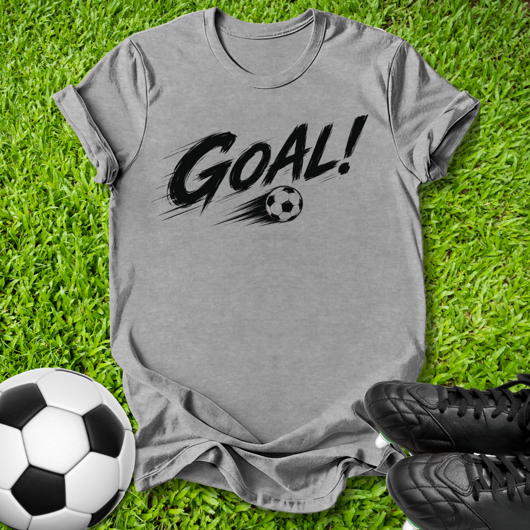 A heather gray short-sleeve t-shirt with a black 'GOAL!' graphic and soccer ball print, laid flat on green grass with a soccer ball and black cleats as props.