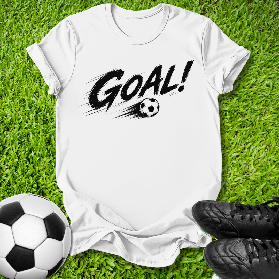 A white short-sleeve T-shirt laid flat on green grass, featuring a black 'GOAL!' graphic with a soccer ball motif. A black-and-white soccer ball sits at the lower left, and black cleats are at the lower right.