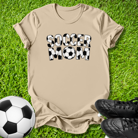 A beige graphic t-shirt laid flat on green grass, featuring the words 'SOCCER MOM' in black and white soccer-ball-patterned letters. A soccer ball is visible in the bottom-left corner and a pair of black cleats is visible in the bottom-right corner.