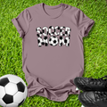 A mauve short-sleeve t-shirt with 'SOCCER MOM' printed in black-and-white soccer-ball patterned letters, laid flat on green grass with a soccer ball and black cleats nearby.