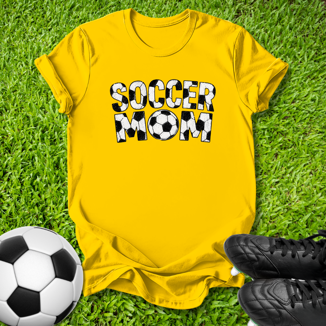 A bright yellow t-shirt with 'SOCCER MOM' printed in bold black and white soccer-ball-patterned letters, laid flat on green grass with a soccer ball and black cleats nearby.