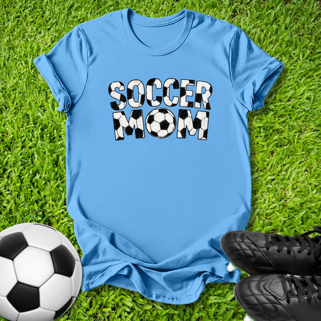 A light blue short-sleeve t-shirt laid flat on green grass, featuring the words 'SOCCER MOM' in bold black-and-white soccer-ball patterned letters. A soccer ball and black cleats are placed around it as styling props.
