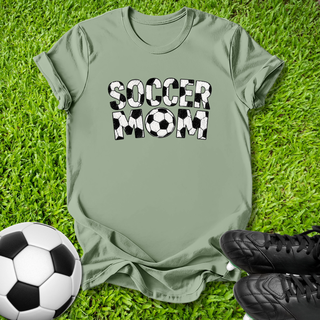 An olive green t-shirt with 'SOCCER MOM' printed in black and white soccer-ball-patterned letters, laid flat on green grass with a soccer ball and black cleats as props.