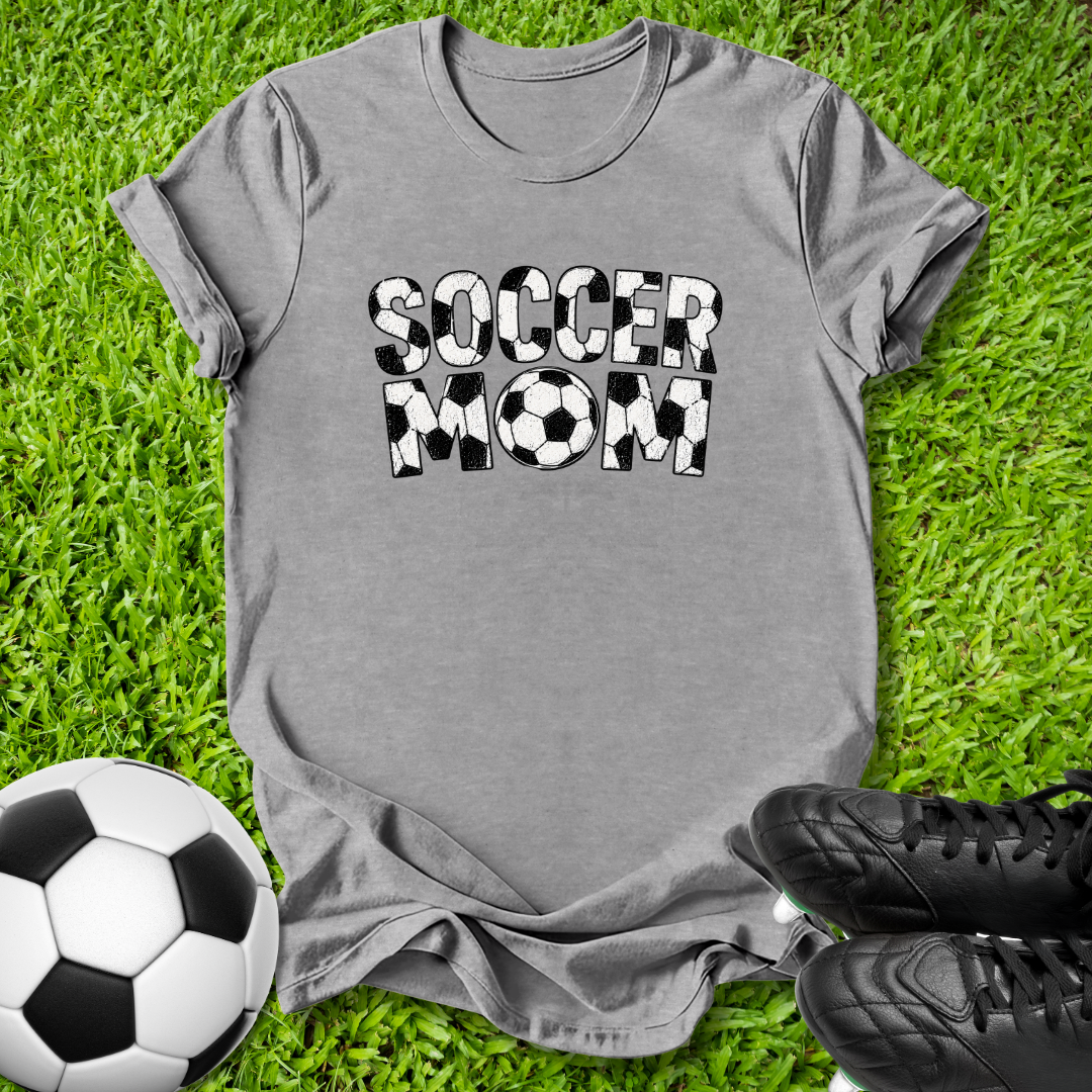 A gray short-sleeve t-shirt with 'SOCCER MOM' printed in black and white soccer-ball-patterned letters, laid flat on green grass with a soccer ball and black cleats nearby.