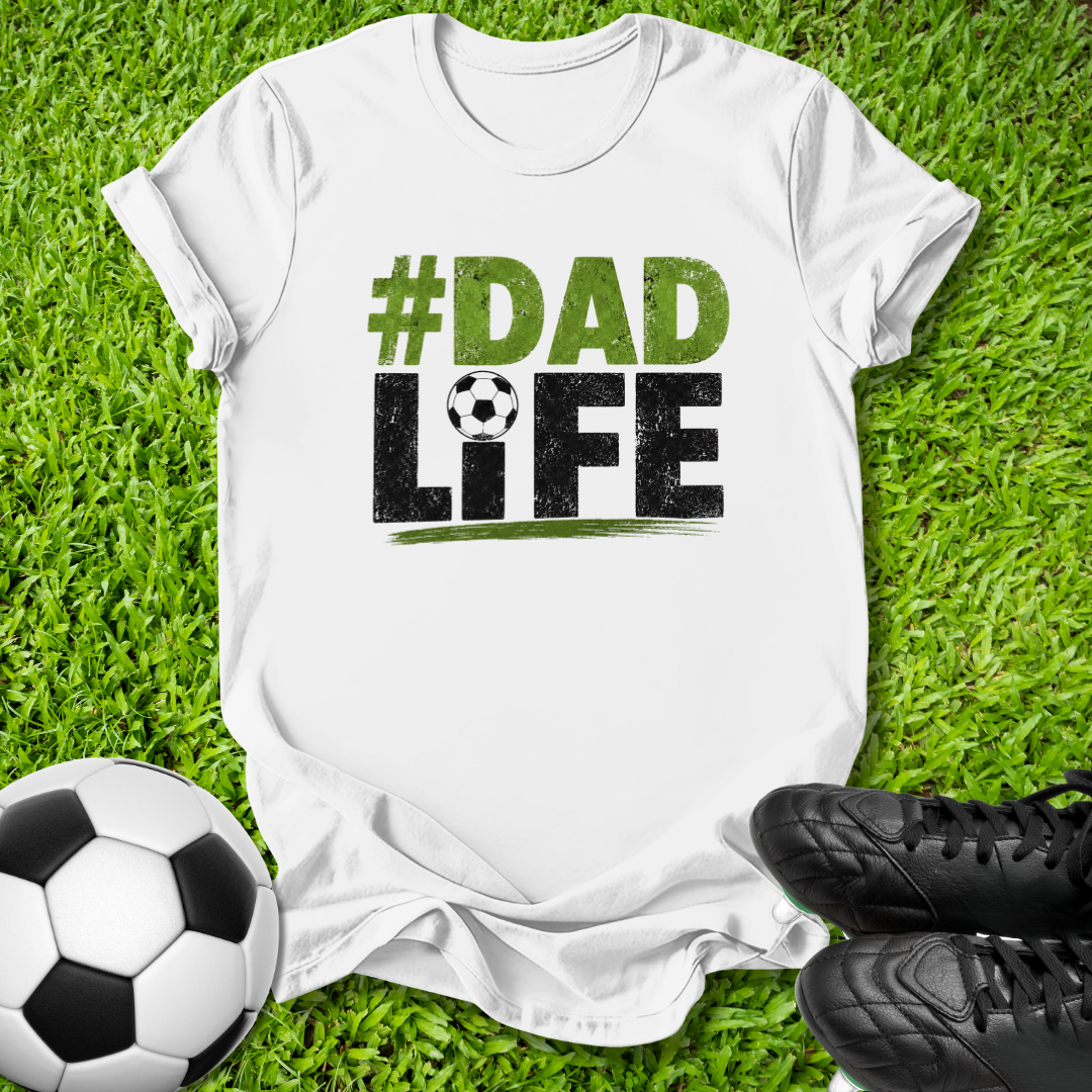 A white short-sleeve T-shirt laid flat on green grass with soccer-themed props, including a soccer ball, black cleats, and a large printed graphic reading “#DAD LIFE” in green and black with a small soccer ball motif.