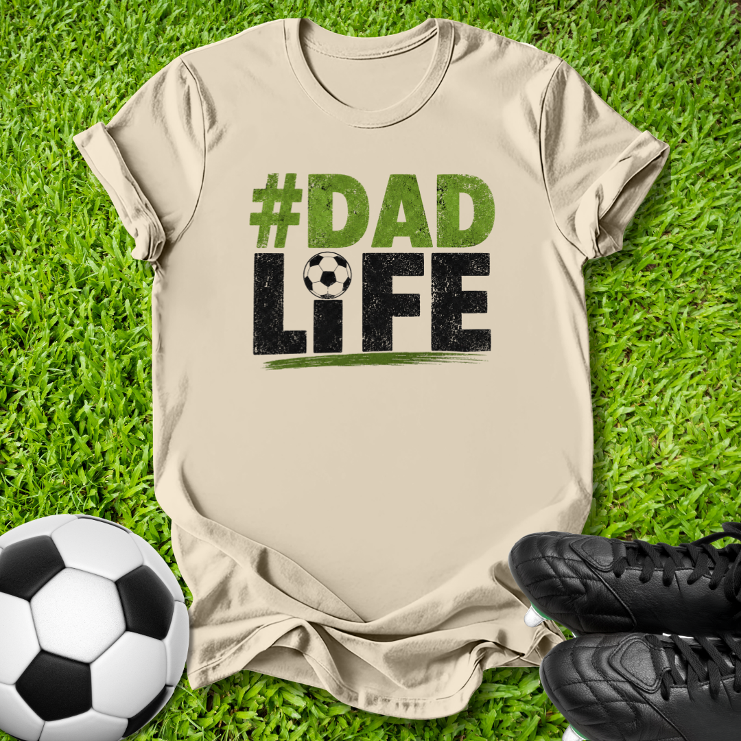 A beige graphic t-shirt laid flat on green grass, featuring the text '#DAD LIFE' with a soccer ball graphic. A soccer ball and black cleats are placed around it as props.