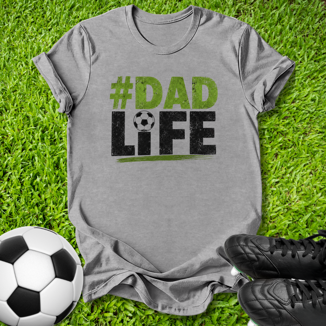 A heather gray graphic t-shirt laid flat on green grass, featuring the text '#DAD LIFE' in distressed green and black lettering with a soccer ball motif. A soccer ball and black cleats are styled around the shirt.