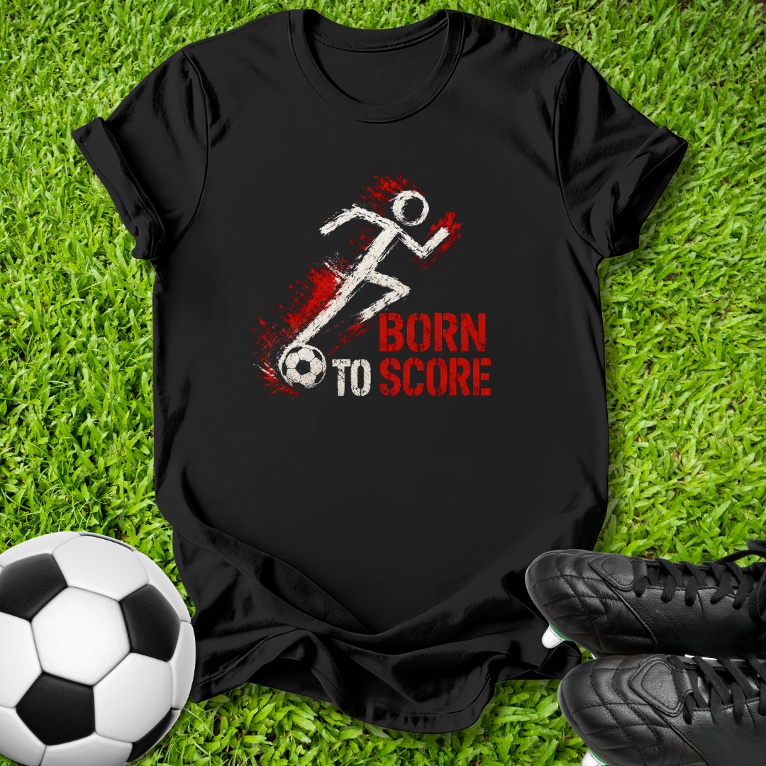 A black short-sleeve graphic T-shirt laid flat on green grass, with a soccer-themed print showing a white running stick figure, a soccer ball, and red text reading 'BORN TO SCORE.' A soccer ball and black cleats are placed around it as styling props.