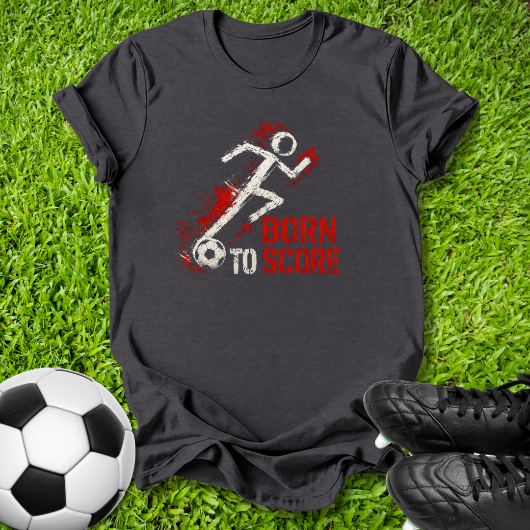 A black short-sleeve graphic t-shirt laid flat on green grass, featuring a white stick figure running with a soccer ball and red text that reads 'BORN TO SCORE'. A soccer ball and black cleats are placed around it as props.