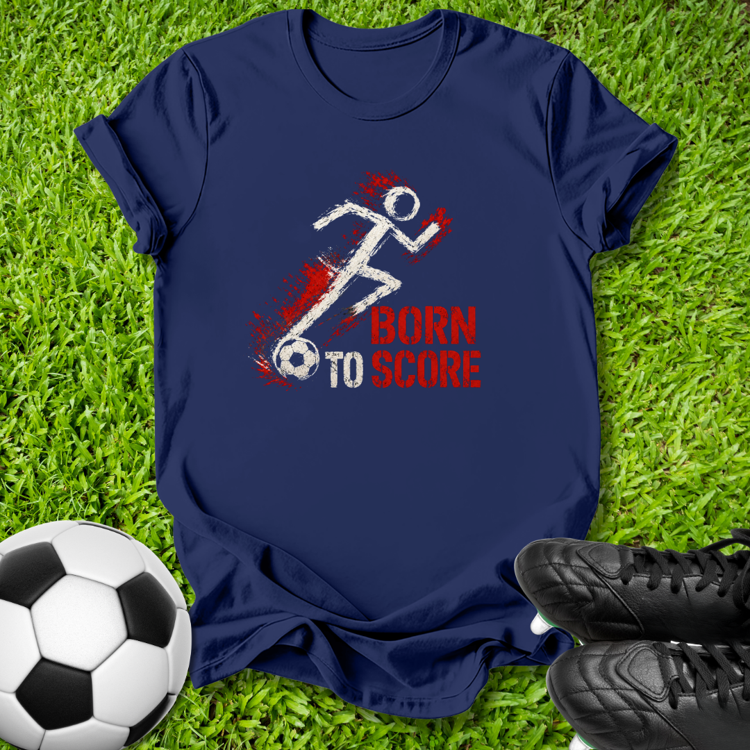 A navy blue short-sleeve t-shirt with a 'BORN TO SCORE' graphic, laid flat on green grass with a soccer ball and black cleats as props.