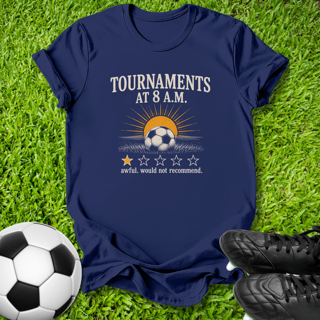 A navy blue graphic t-shirt laid flat on green grass, featuring a soccer-themed design with the text 'TOURNAMENTS AT 8 A.M.