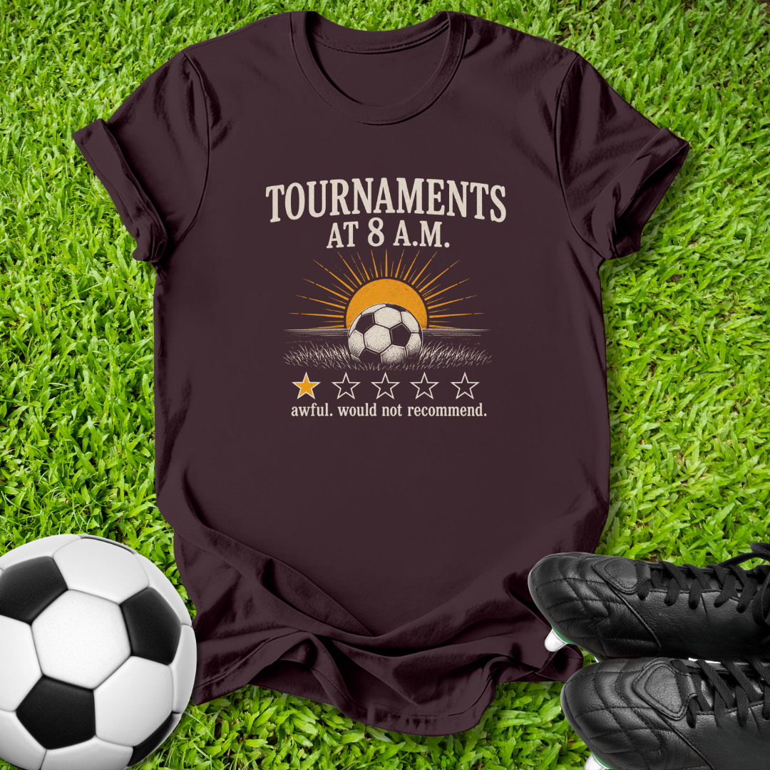 A dark brown graphic t-shirt laid flat on green grass, featuring a soccer-themed design with the text 'TOURNAMENTS AT 8 A.M.
