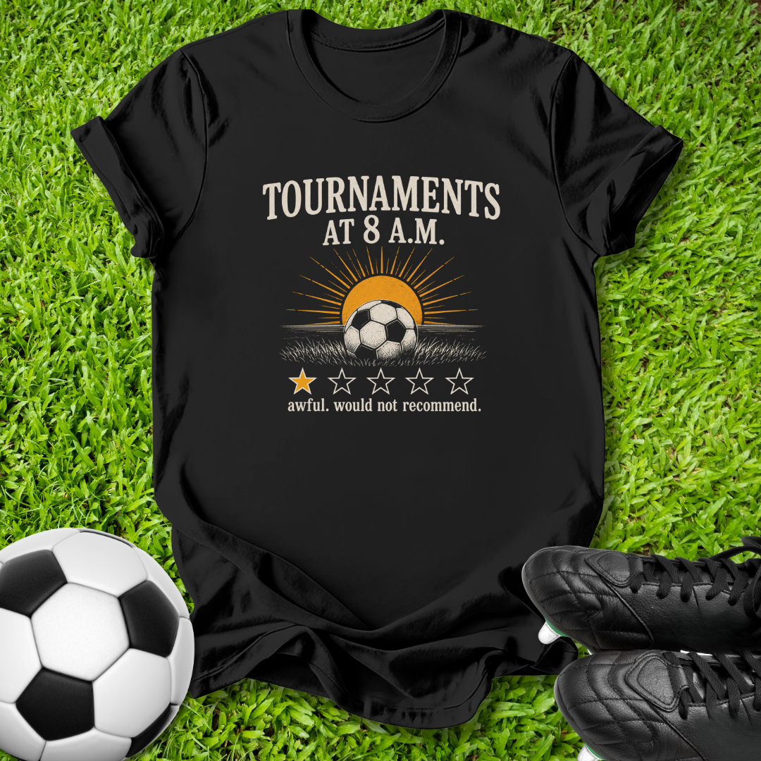 A black graphic t-shirt laid flat on green grass, featuring a soccer-themed design with the text 'TOURNAMENTS AT 8 A.M.