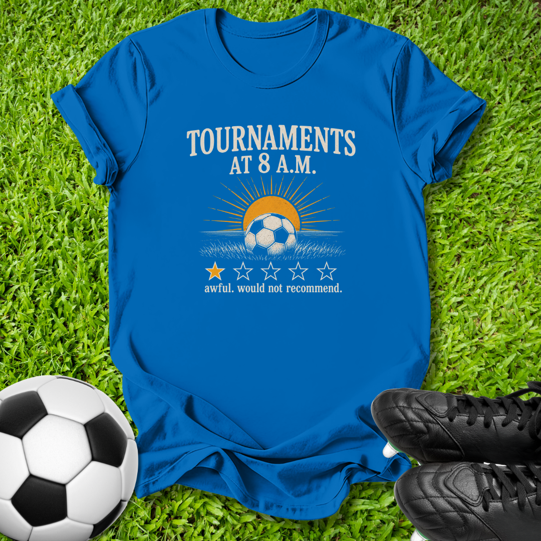 A royal blue graphic t-shirt laid flat on green grass, featuring a soccer-themed design with the text 'TOURNAMENTS AT 8 A.M.