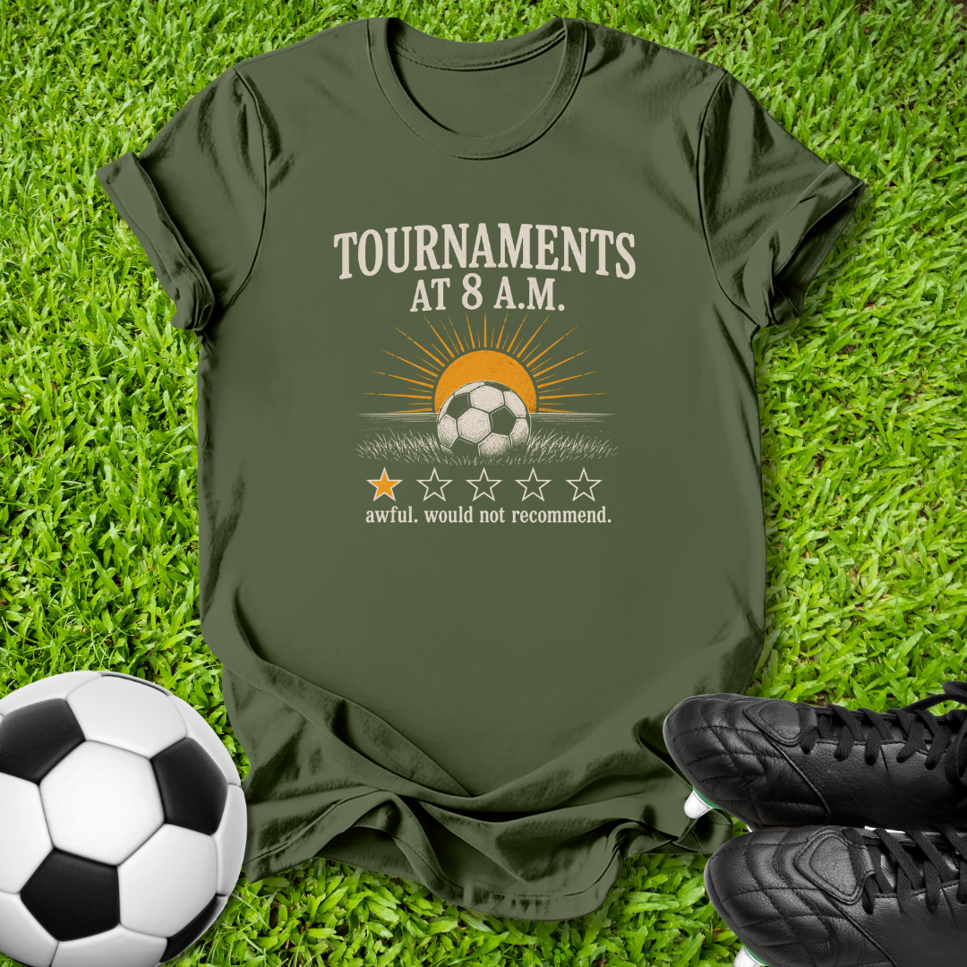 An olive green graphic t-shirt laid flat on green grass, styled with a soccer ball on the bottom left and black cleats on the bottom right. The t-shirt features a graphic of a soccer ball under a rising sun with the text 'TOURNAMENTS AT 8 A.