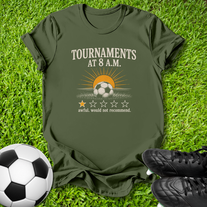 An olive green graphic t-shirt laid flat on green grass, styled with a soccer ball on the bottom left and black cleats on the bottom right. The t-shirt features a graphic of a soccer ball under a rising sun with the text 'TOURNAMENTS AT 8 A.