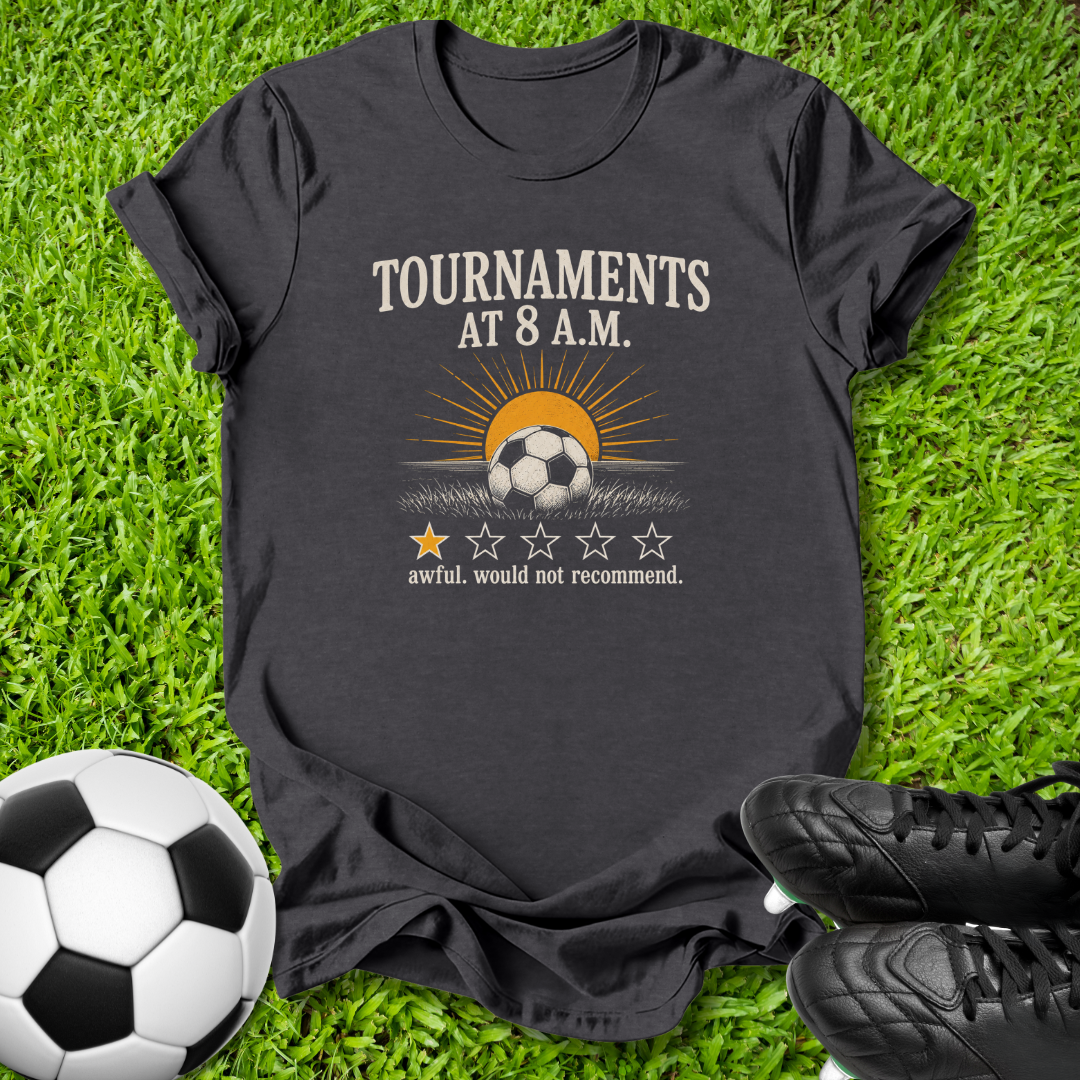 A dark charcoal gray graphic t-shirt laid flat on green grass, styled with a soccer ball on the left and black cleats on the right. The shirt features a graphic of a soccer ball under a rising sun with the text 'TOURNAMENTS AT 8 A.