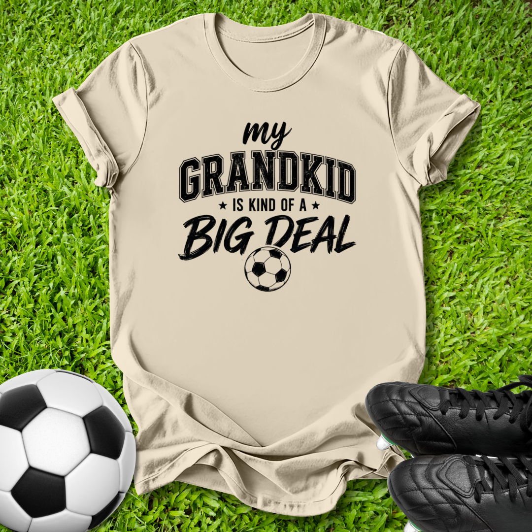 A beige graphic t-shirt laid flat on green grass, featuring the text 'my GRANDKID IS KIND OF A BIG DEAL' with a soccer ball graphic. A soccer ball and black cleats are styled around the shirt.