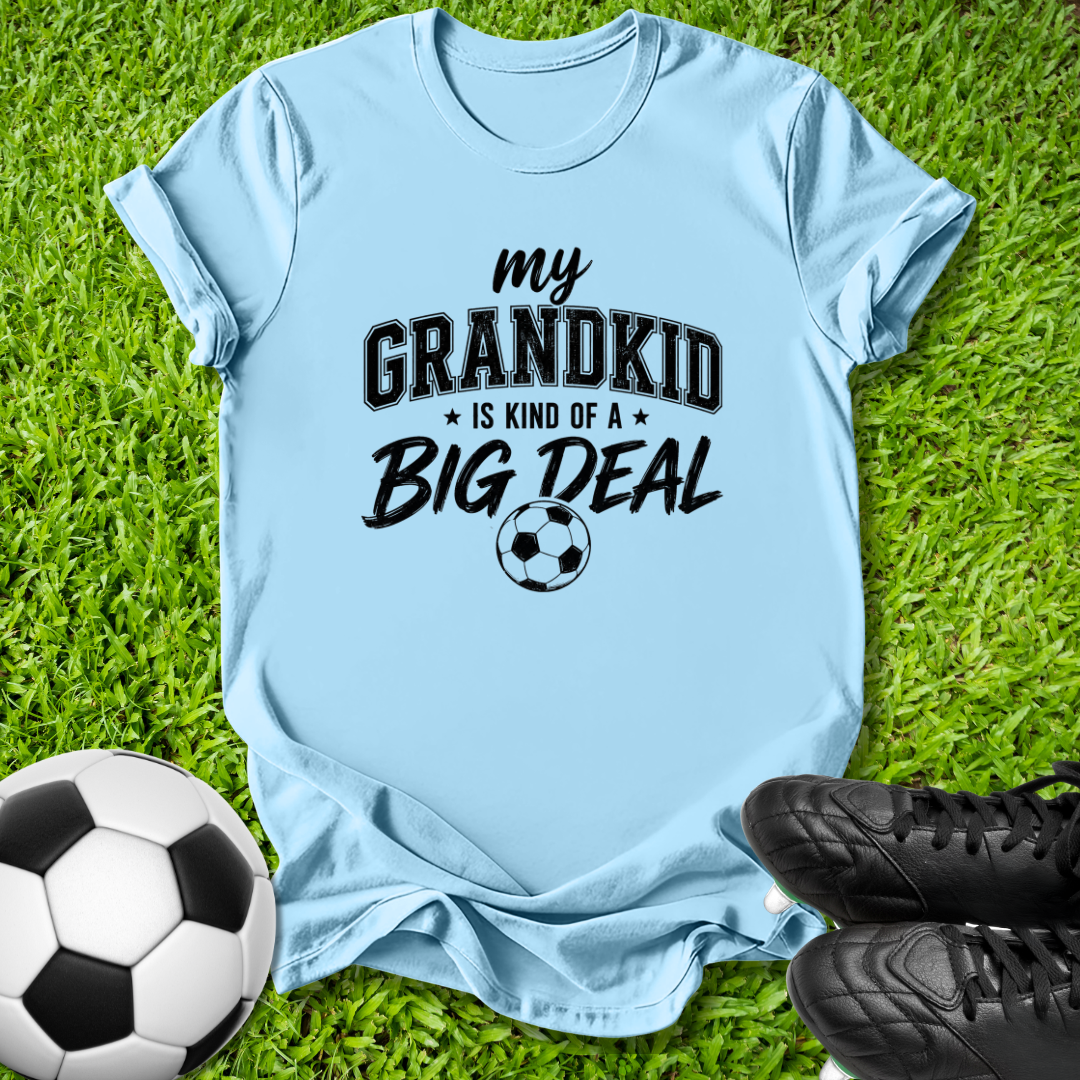 A light blue graphic t-shirt laid flat on green grass, featuring the text 'my GRANDKID IS KIND OF A BIG DEAL' with a soccer ball graphic. A soccer ball and black cleats are placed around it as props.