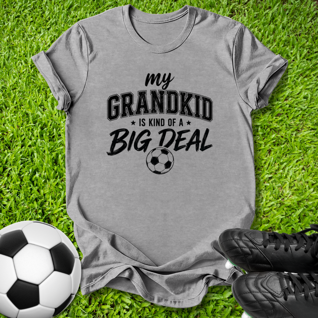A heather gray short-sleeve t-shirt with a black graphic text reading 'my GRANDKID IS KIND OF A BIG DEAL' and a soccer ball icon, laid flat on green grass with a soccer ball and black cleats nearby.