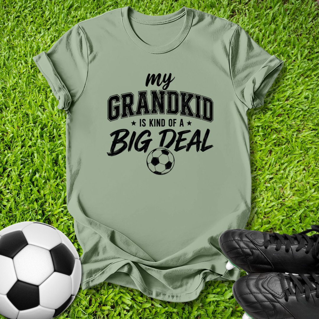 An olive green graphic t-shirt laid flat on green grass, featuring the text 'my GRANDKID IS KIND OF A BIG DEAL' with a soccer ball graphic. A soccer ball and black cleats are styled as props around the shirt.