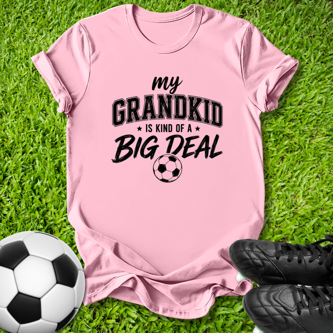 A light pink short-sleeve t-shirt with a black graphic text reading 'my GRANDKID IS KIND OF A BIG DEAL' and a soccer ball graphic, laid flat on green grass with a soccer ball and black cleats as props.