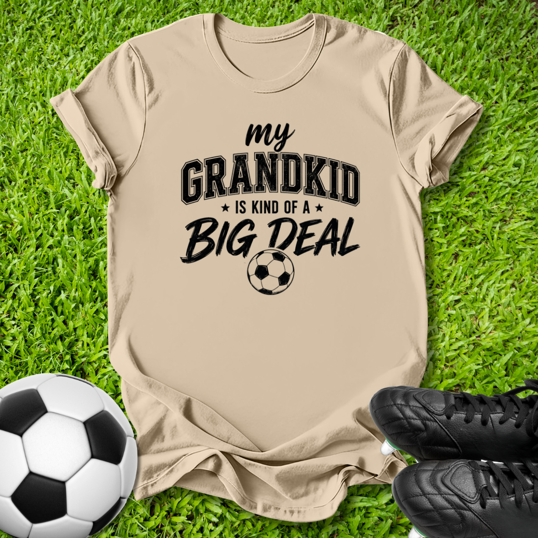 A beige graphic t-shirt laid flat on green grass, featuring the text 'my GRANDKID IS KIND OF A BIG DEAL' with a soccer ball graphic. A soccer ball and black cleats are placed around it as props.