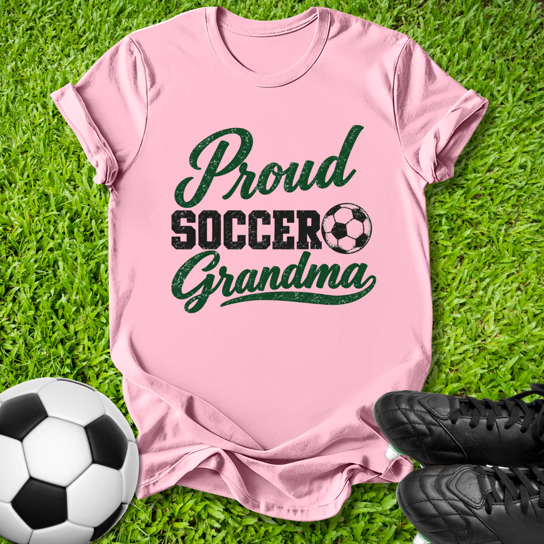 A pink short-sleeve t-shirt with a 'Proud Soccer Grandma' graphic in dark green and black, laid flat on green grass with a soccer ball and black cleats as props.