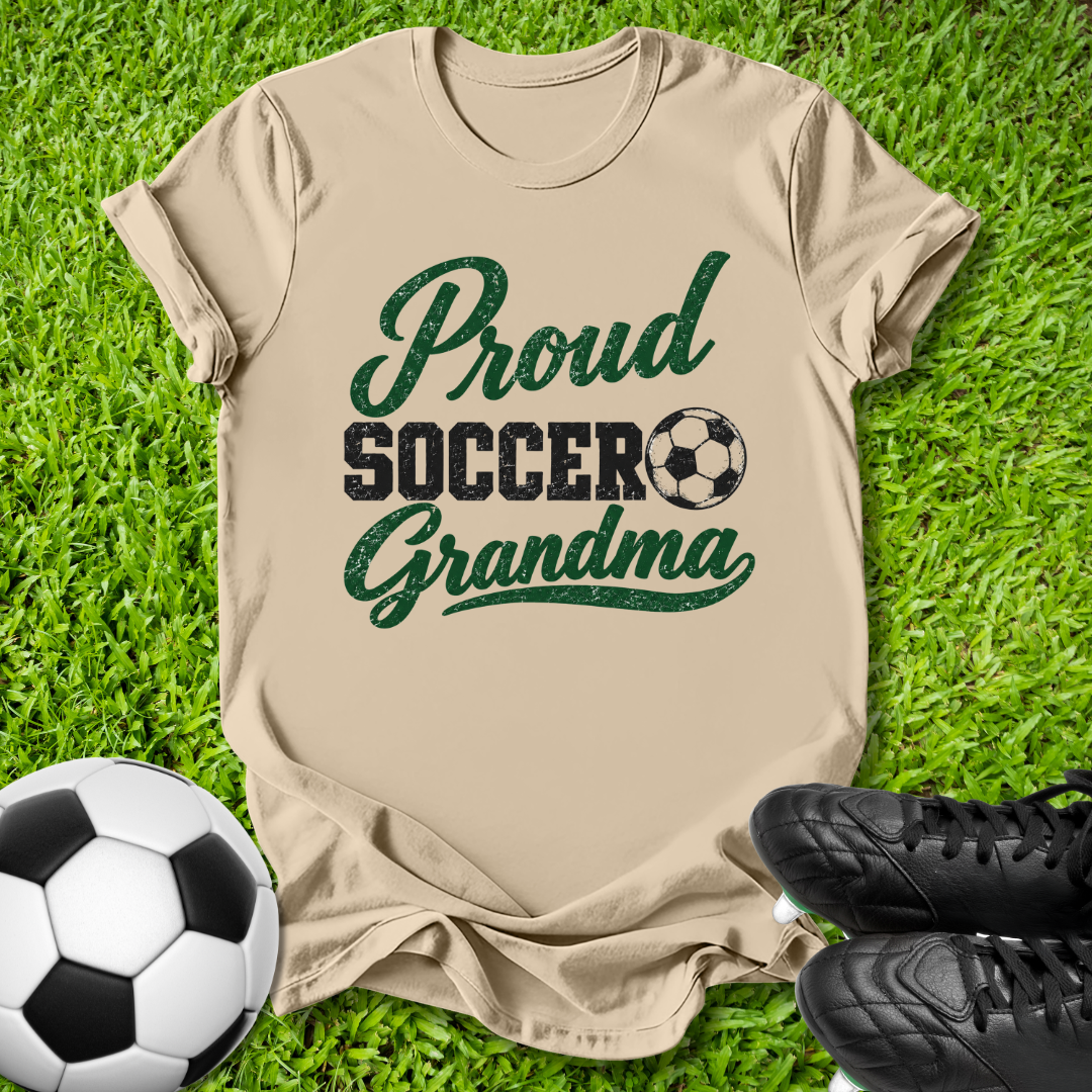 A beige short-sleeve t-shirt laid flat on green grass, printed with the words 'Proud Soccer Grandma' in dark green and black, alongside a soccer ball graphic. A soccer ball and black cleats are placed around it as props.