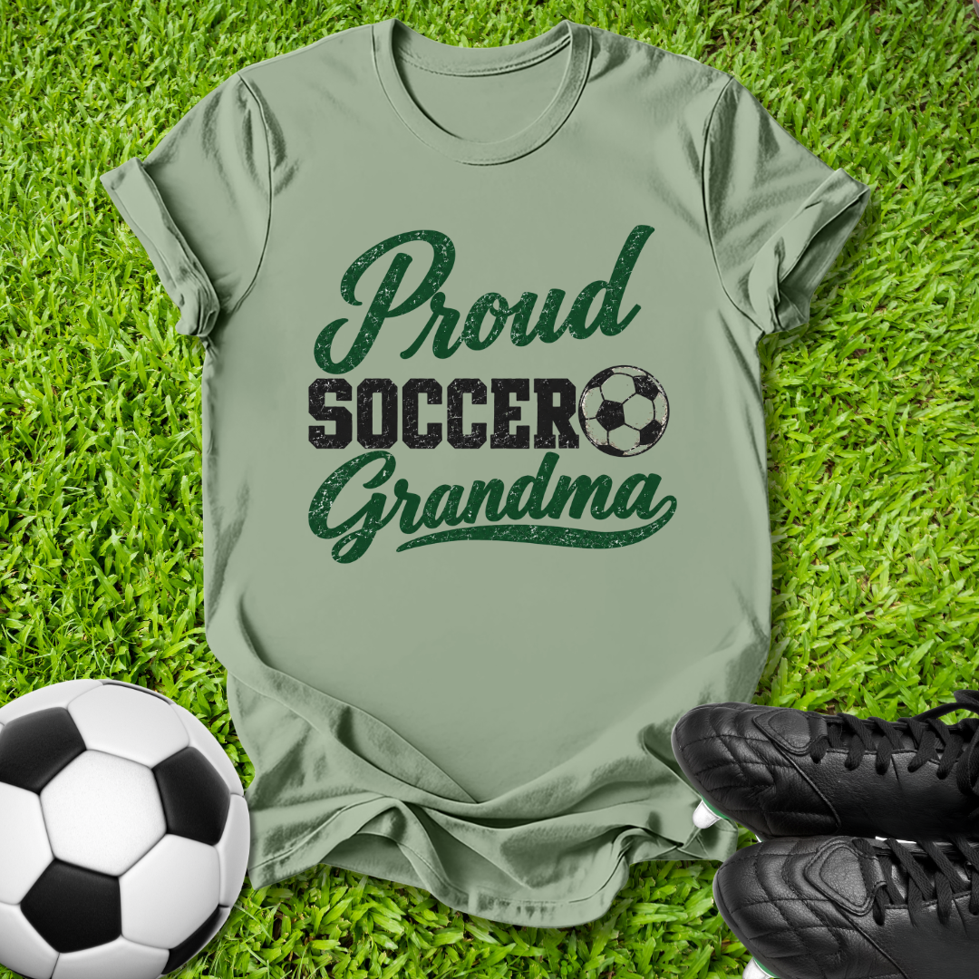 An olive green t-shirt with a 'Proud Soccer Grandma' graphic printed on the front, laid flat on green grass with a soccer ball and black cleats as props.