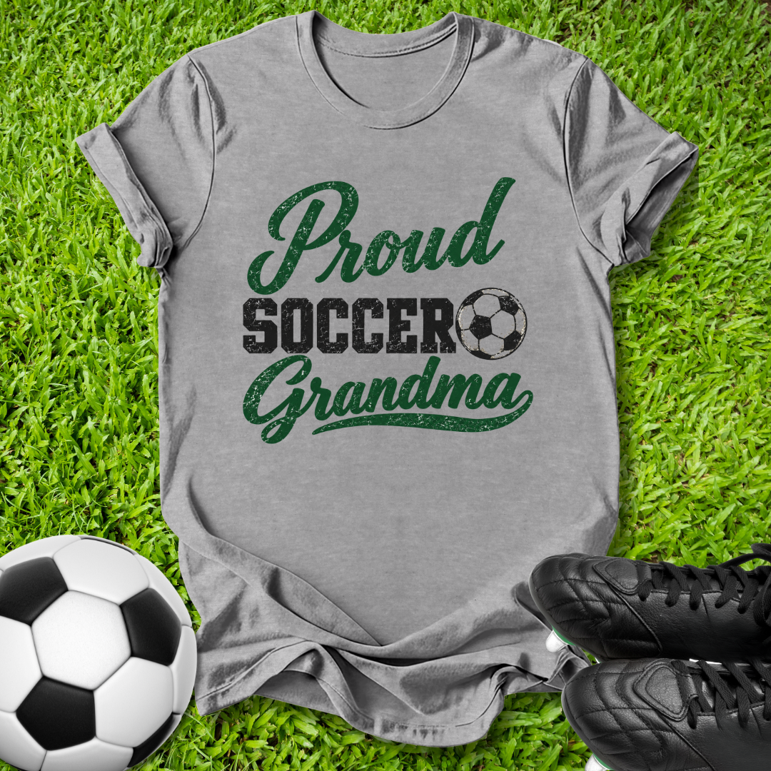 A heather gray short-sleeve t-shirt with a distressed green and black graphic reading 'Proud Soccer Grandma' with a soccer ball icon, laid flat on green grass with a soccer ball and black cleats nearby.