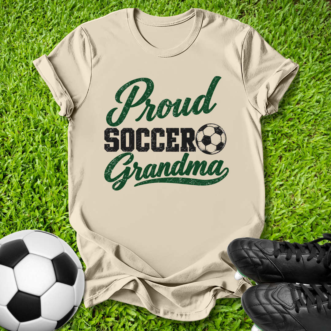 A beige short-sleeve t-shirt laid flat on green grass, featuring the text 'Proud Soccer Grandma' in dark green and black, with a soccer ball graphic. A soccer ball and black cleats are placed around it as props.