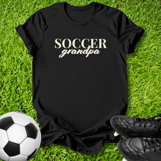 A black short-sleeve t-shirt with 'SOCCER grandpa' printed in white across the chest, laid flat on green grass with a soccer ball and black cleats nearby.