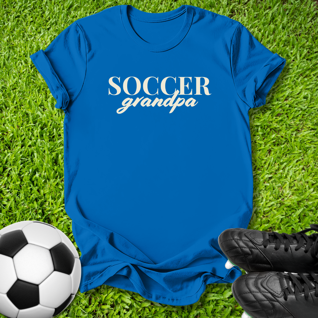 A royal blue short-sleeve t-shirt with 'SOCCER grandpa' printed in white across the chest, laid flat on green grass with a soccer ball and black cleats nearby.