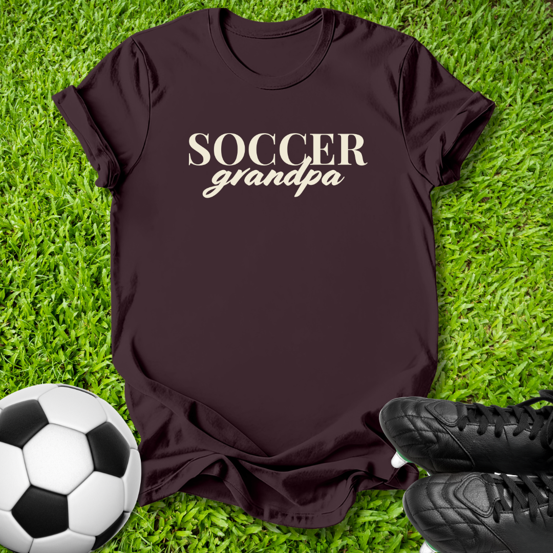 A dark brown short-sleeve t-shirt with 'SOCCER grandpa' printed in white across the chest, laid flat on green grass with a soccer ball and black cleats as props.