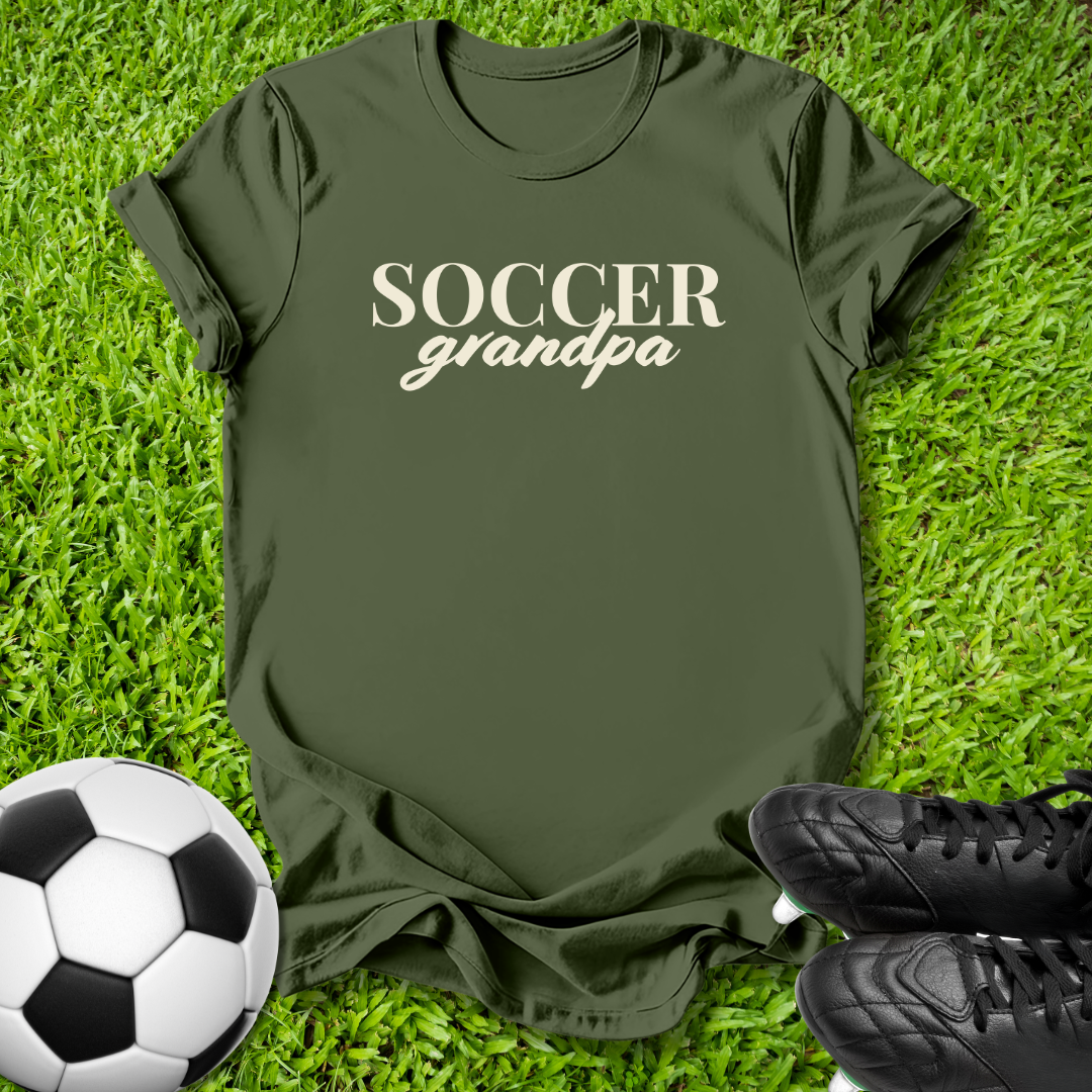An olive green t-shirt with 'SOCCER grandpa' printed in white across the chest, laid flat on green grass with a soccer ball and black cleats as props.
