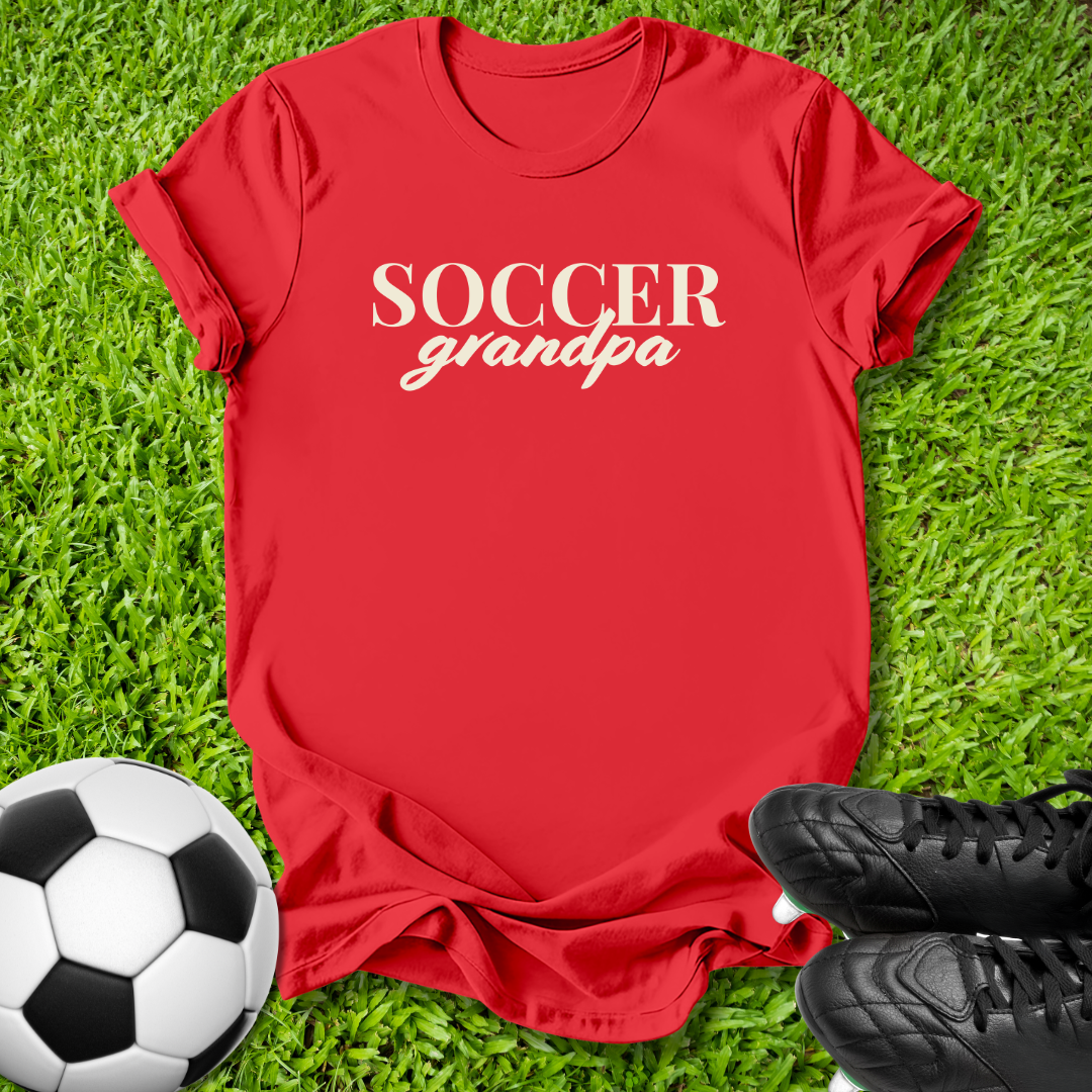 A red short-sleeve t-shirt with 'SOCCER grandpa' printed in white across the chest, laid flat on green grass with a soccer ball and black cleats nearby.
