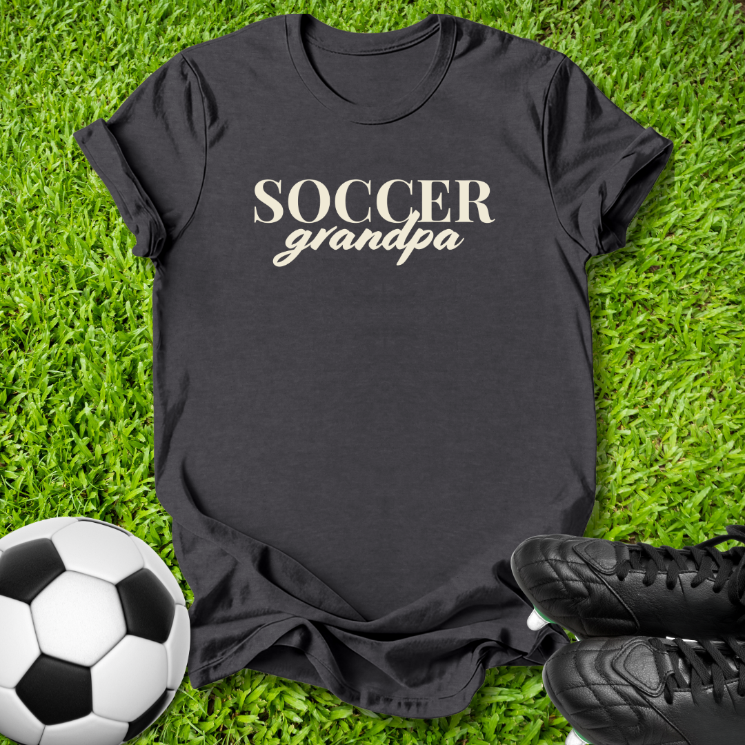 A black short-sleeve t-shirt with 'SOCCER grandpa' printed in beige across the chest, laid flat on green grass with a soccer ball and black cleats nearby.