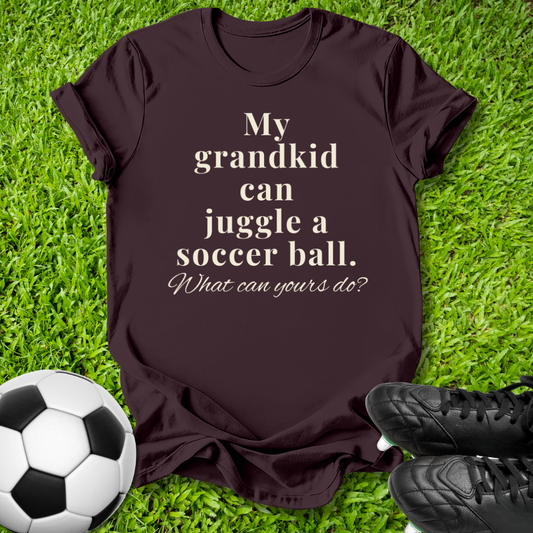 A dark brown short-sleeve t-shirt with the text 'My grandkid can juggle a soccer ball. What can yours do?' printed in beige, laid flat on green grass with a soccer ball and black cleats as props.
