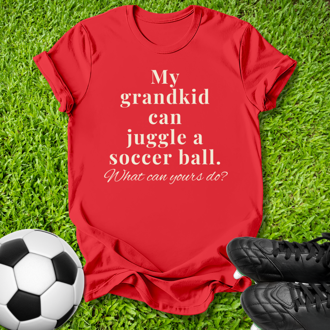 A red short-sleeve t-shirt with white text reading 'My grandkid can juggle a soccer ball. What can yours do?' laid flat on green grass, styled with a soccer ball and black cleats nearby.
