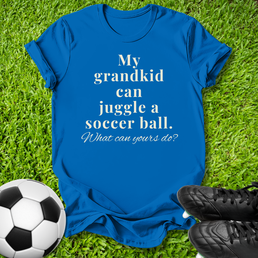 A royal blue graphic t-shirt laid flat on green grass, featuring the white printed text 'My grandkid can juggle a soccer ball. What can yours do?'.
