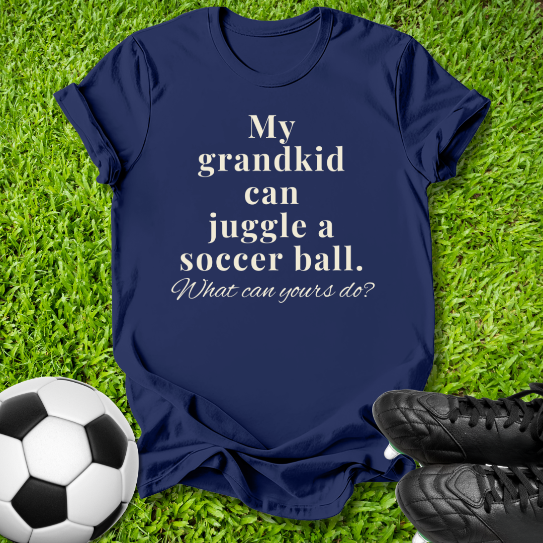 A navy blue graphic t-shirt laid flat on green grass, featuring the white printed text 'My grandkid can juggle a soccer ball. What can yours do?'.