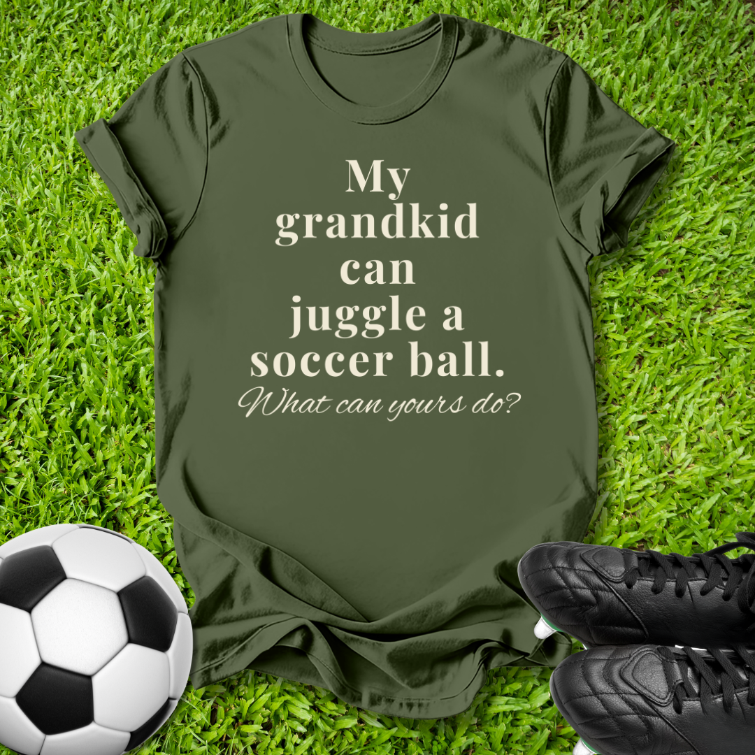 An olive green t-shirt with the text 'My grandkid can juggle a soccer ball. What can yours do?' printed in cream, laid flat on green grass with a soccer ball and black cleats as props.