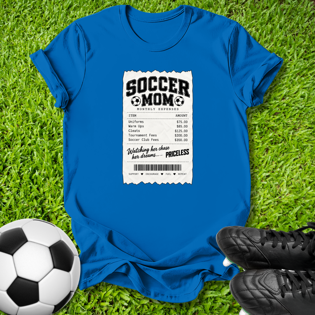 A bright blue graphic t-shirt laid flat on green grass, featuring a white and black 'Soccer Mom Receipt' print on the front. A soccer ball is on the bottom left, and black soccer cleats are on the bottom right.