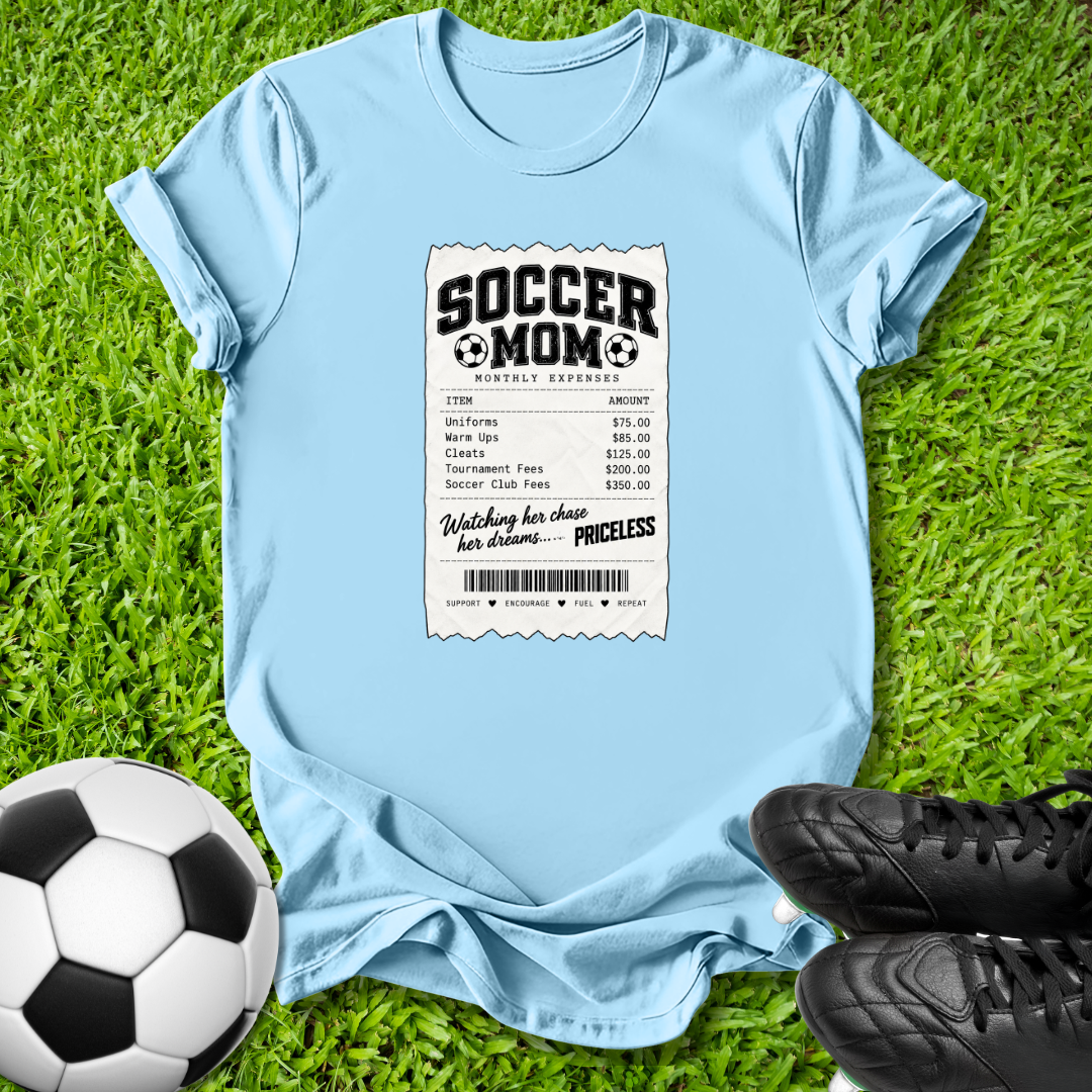A light blue graphic t-shirt laid flat on green grass, styled with a soccer ball on the left and black soccer cleats on the right. The t-shirt features a white and black 'SOCCER MOM' receipt print graphic on the front.