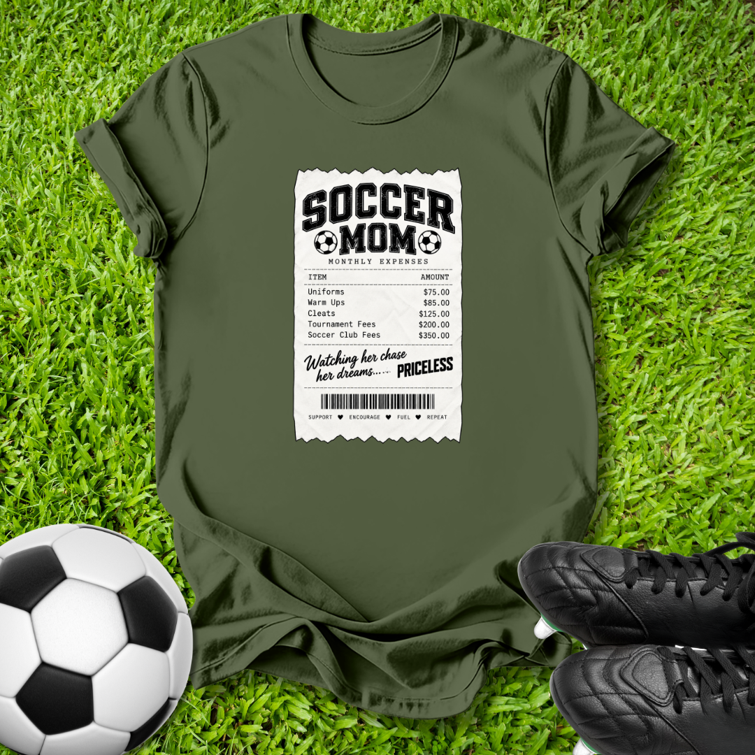 An olive green short-sleeve t-shirt laid flat on green grass, featuring a white and black 'Soccer Mom Receipt' graphic print on the front. A black and white soccer ball is on the bottom left, and black soccer cleats are on the bottom right.