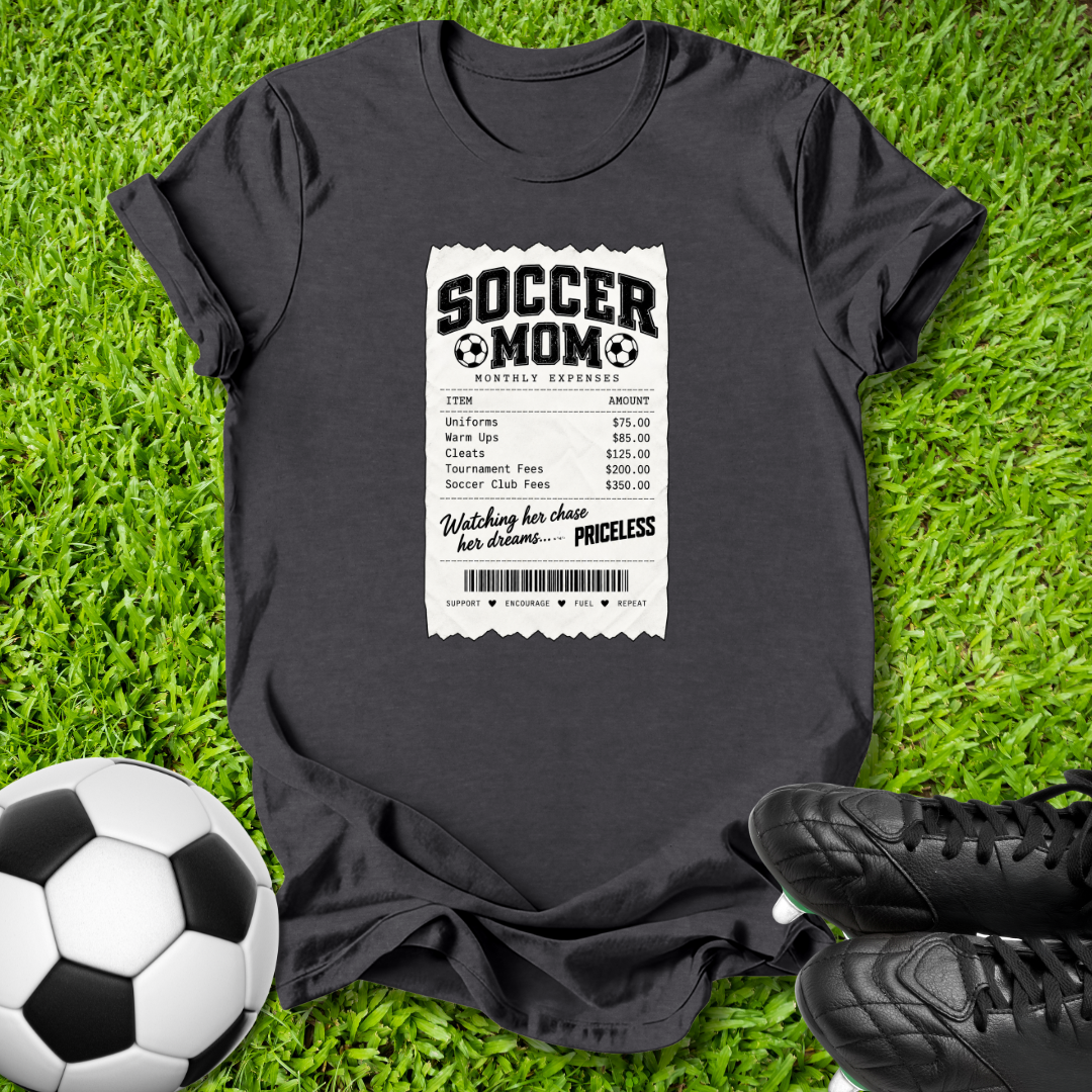 A dark charcoal gray graphic t-shirt laid flat on green grass, styled with a soccer ball on the left and black cleats on the right. The shirt features a white and black 'Soccer Mom' receipt print graphic on the front.