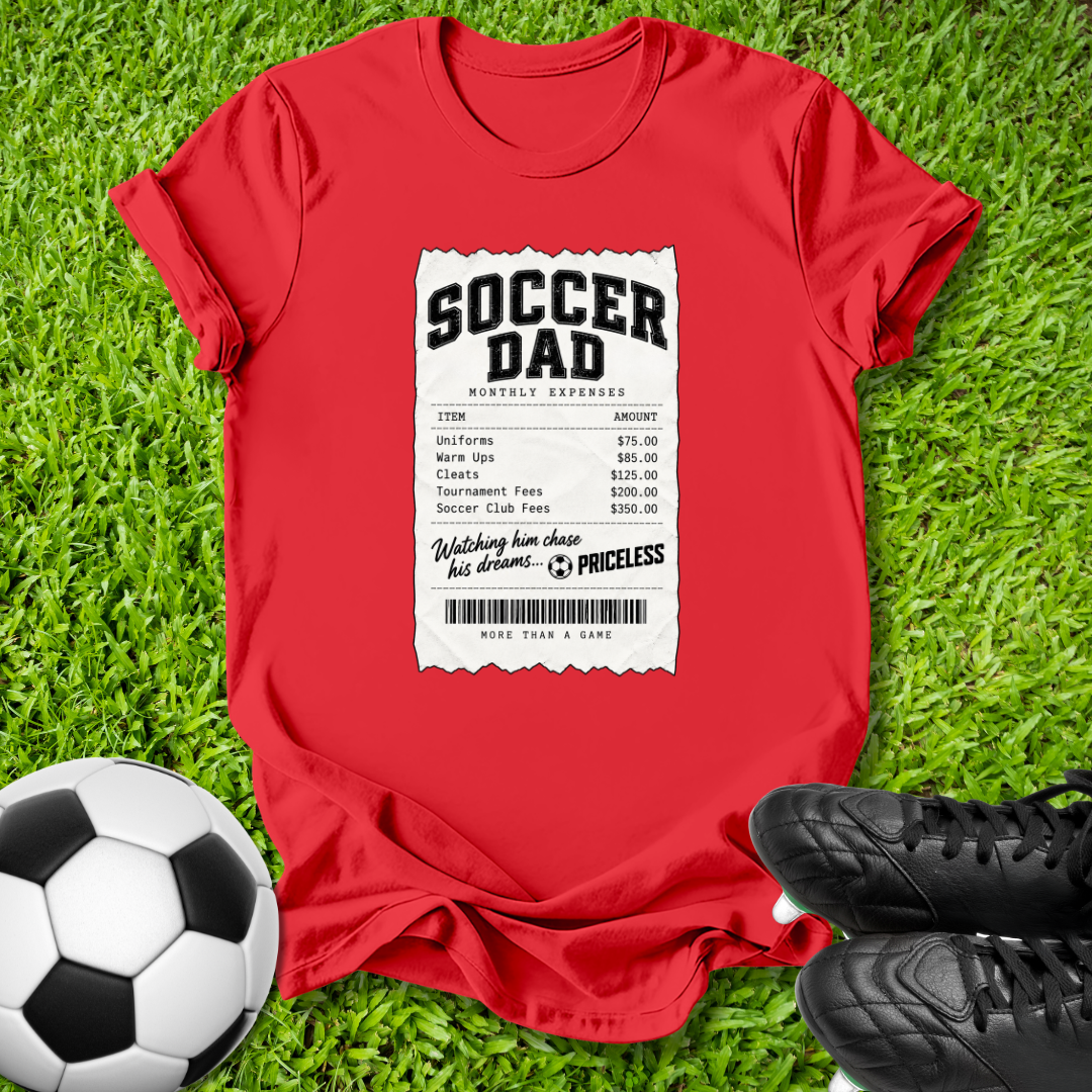 A red short-sleeve t-shirt laid flat on green grass, featuring a white printed receipt-style graphic that reads 'SOCCER DAD' with soccer-related expenses. A black-and-white soccer ball sits at the lower left, and black cleats are at the lower right.